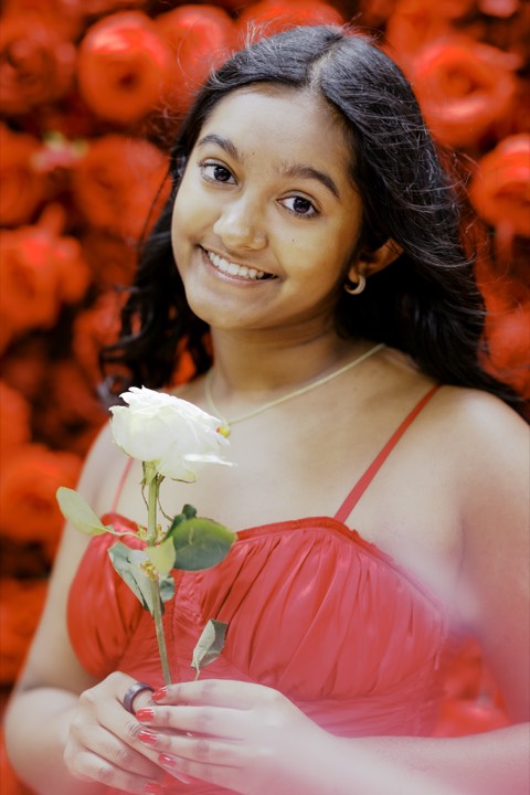 Anya in a red gown holding a single white rose against a wall of red roses, smiling