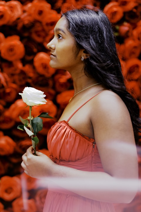 Anya in profile, studying a white rose against the red rose wall